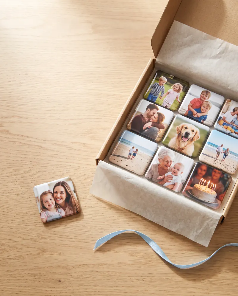 Um ímã com foto de mãe e filha sozinho sobre a mesa, ao lado de uma caixa kraft cheia de ímãs com fotos de família, pets e viagens